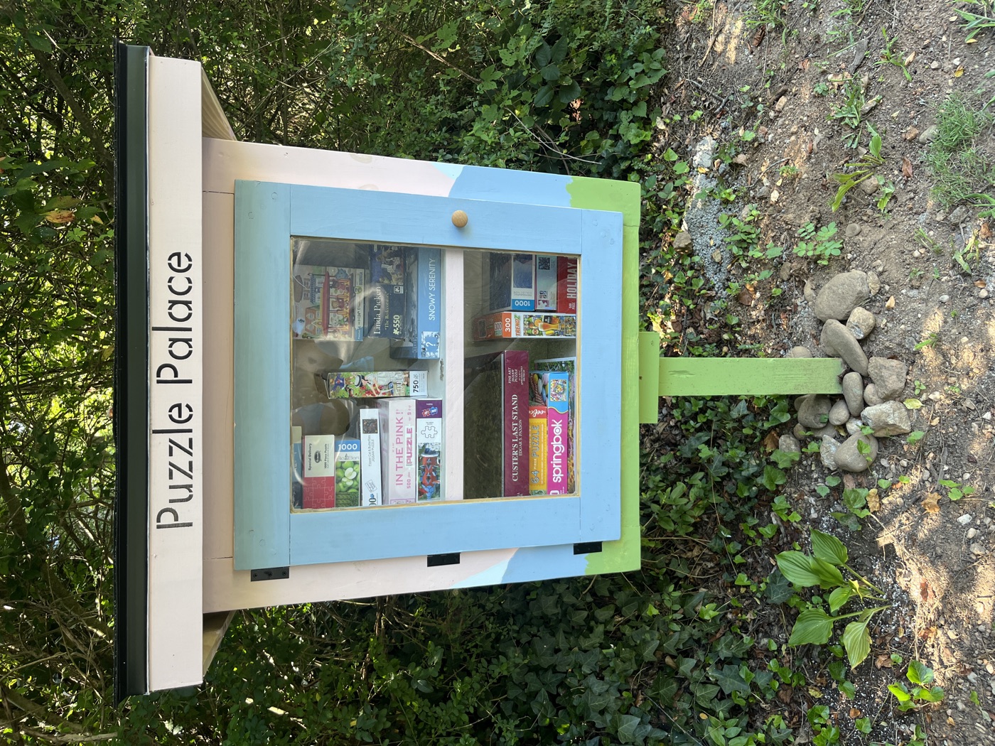 The Puzzle Palace: a pink box with a dark roof, a stencil-cut Puzzle Palace sign, and a periwinkle glass door over two shelves of puzzle boxes.